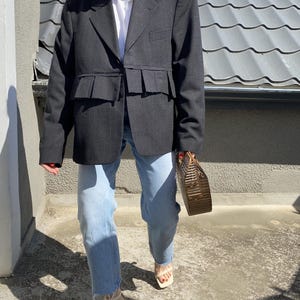 Könnte beinhalten: Ein grauer Blazer mit einem Rüschen-Saum und einem weißen Hemd mit Kragen darunter. Der Blazer wird mit hellblauen Jeans und weißen Sandaletten mit Absatz getragen.