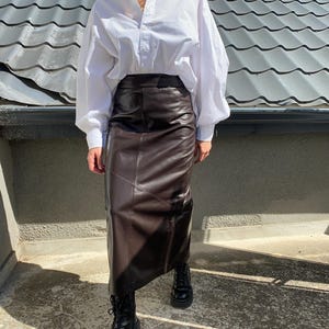 Op de afbeelding: Een witte blouse met volumineuze mouwen gecombineerd met een donkerbruine leren rok. De outfit wordt gecompleteerd met zwarte combat boots. Het ensemble is vastgelegd tegen de achtergrond van een pannendak en een heldere blauwe lucht.