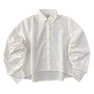 Puede incluir: Una camisa blanca con cuello clásico y mangas largas. Las mangas están recogidas en los puños, creando un efecto voluminoso y fruncido. La camisa tiene un dobladillo curvo y un diseño minimalista.