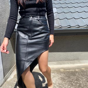 Könnte beinhalten: Schwarzer Lederrock mit asymmetrischem Saum. Der Rock wird mit einem schwarzen Langarm-Oberteil kombiniert. Das Outfit wird durch beige spitz zulaufende Absätze ergänzt. Der Rock hat ein klassisches, elegantes Design.