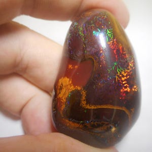 Könnte beinhalten: Ein glatter, polierter, tropfenförmiger Opal-Edelstein mit einer dunkelbraunen, roten und violetten Basis. Der Stein hat irisierende Blitze von Grün, Blau und Orange.