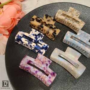 May include: Six different patterned hair clips. The clips are made of plastic and have a curved shape. The patterns include blue and white, brown and white, purple and white, and iridescent.