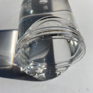 May include: A clear plastic container with a textured surface. The container is filled with a clear liquid.