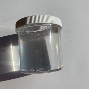May include: A clear plastic jar with a white lid filled with a clear liquid. The liquid has a slight shimmer to it.