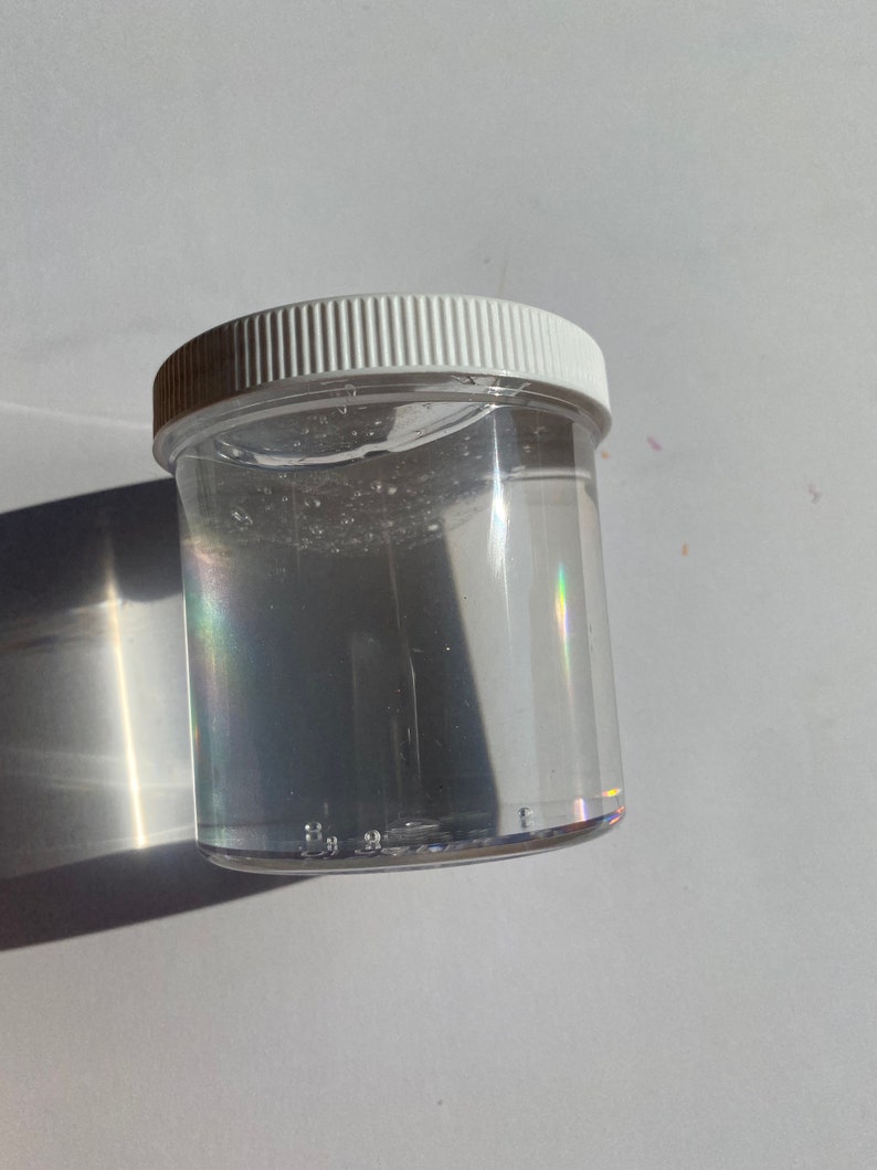 May include: A clear plastic jar with a white lid filled with a clear liquid. The liquid has a slight shimmer to it.