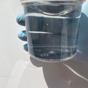 May include: A clear plastic container with a white lid, filled with a clear liquid. The container is being held by a gloved hand.