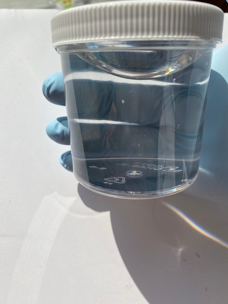 May include: A clear plastic container with a white lid, filled with a clear liquid. The container is being held by a gloved hand.