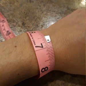 May include: Pink measuring tape wrapped around a wrist, showing a measurement of 7 inches. The tape reads "Made in China".