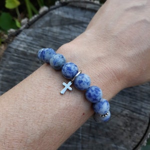 May include: A blue and white gemstone bracelet with a silver cross charm. The bracelet is made of round beads and is worn on a person's wrist.