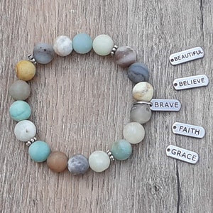 May include: A beaded bracelet with various earth-toned stones, including yellow, blue, and brown. The bracelet is accented with small silver beads. Beside the bracelet are silver charms with the words "BEAUTIFUL", "BELIEVE", "BRAVE", "FAITH", and "GRACE".