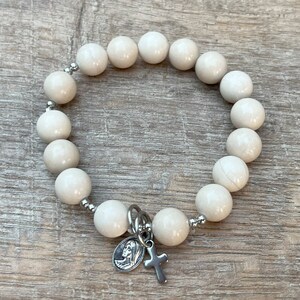 May include: White beaded bracelet with a silver charm featuring a cross and a depiction of the Virgin Mary.