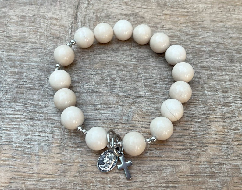 May include: White beaded bracelet with a silver charm featuring a cross and a depiction of the Virgin Mary.