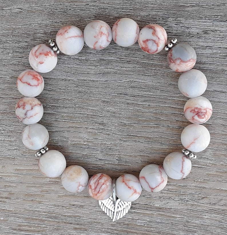 Puede incluir: Un brazalete hecho de cuentas de m&aacute;rmol blanco y rojo con un dije de ala de &aacute;ngel plateado.