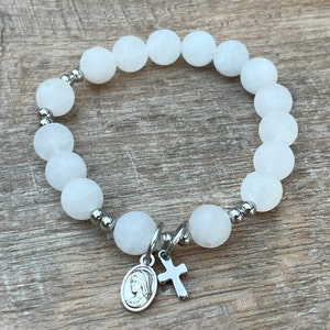 May include: White beaded bracelet with a silver charm featuring a cross and a depiction of the Virgin Mary.