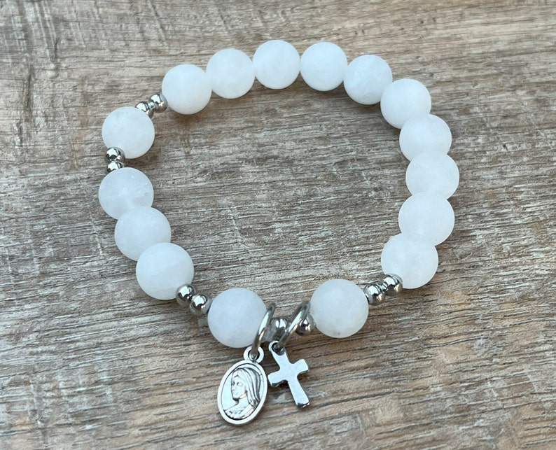 May include: White beaded bracelet with a silver charm featuring a cross and a depiction of the Virgin Mary.
