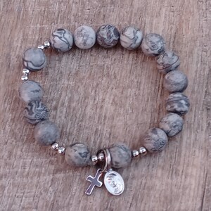 May include: A gray and white stone beaded bracelet with a silver charm featuring a cross and a round medallion.