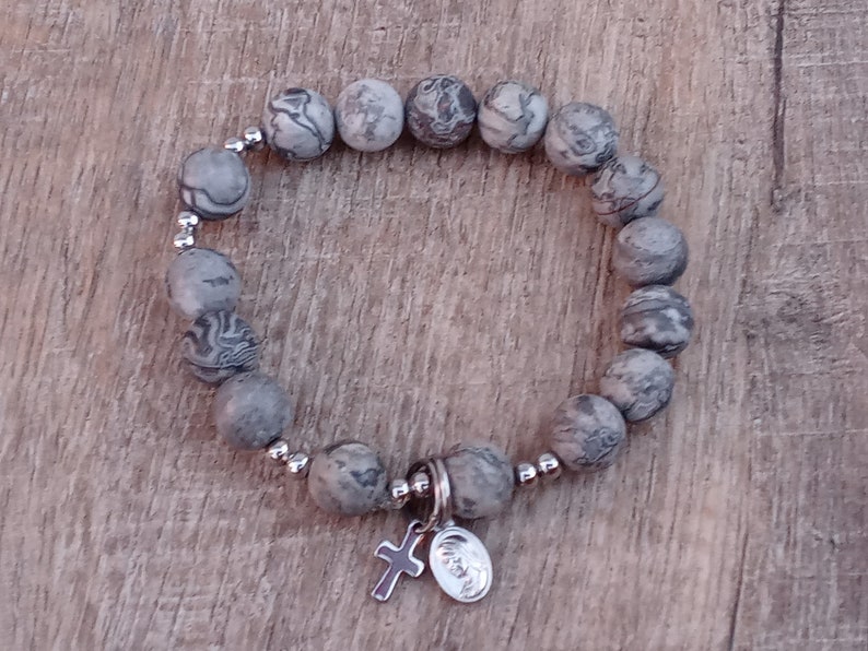 May include: A gray and white stone beaded bracelet with a silver charm featuring a cross and a round medallion.
