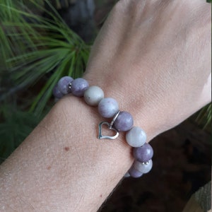 May include: A purple and gray beaded bracelet with a silver heart charm. The bracelet is made of natural stones and is a beautiful piece of jewelry.