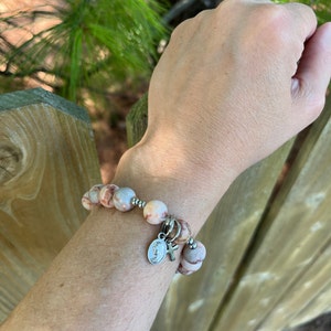 May include: A bracelet made of light brown and white marbled beads with a silver charm featuring a cross and a religious image. The bracelet is worn on a person's wrist.