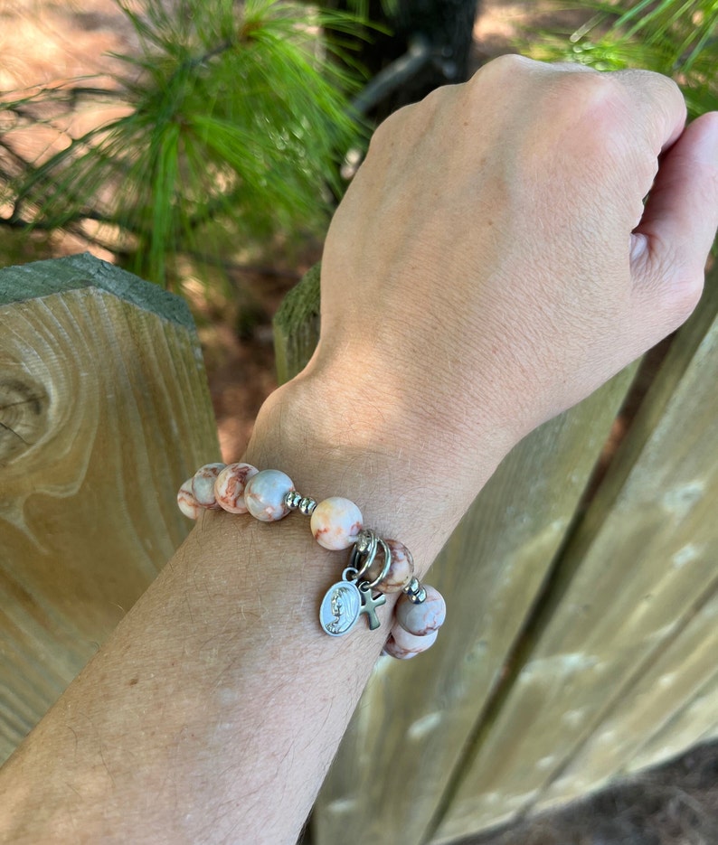 May include: A bracelet made of light brown and white marbled beads with a silver charm featuring a cross and a religious image. The bracelet is worn on a person's wrist.