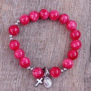 May include: A red beaded bracelet with a silver charm featuring a cross and a depiction of the Virgin Mary.