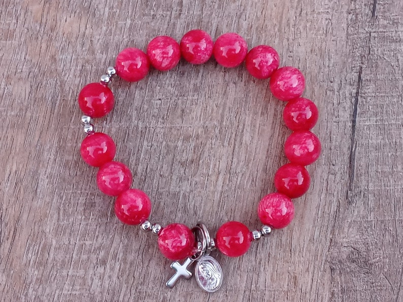 May include: A red beaded bracelet with a silver charm featuring a cross and a depiction of the Virgin Mary.