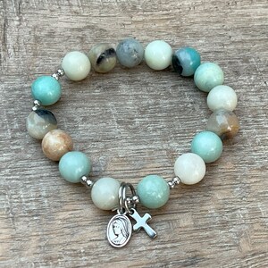 May include: A green and white beaded bracelet with a silver charm featuring a cross and a depiction of the Virgin Mary.