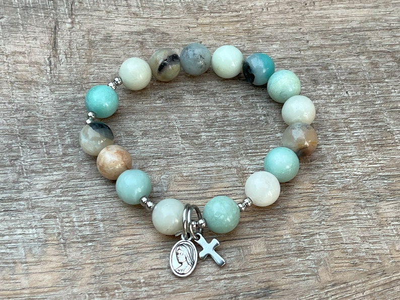 May include: A green and white beaded bracelet with a silver charm featuring a cross and a depiction of the Virgin Mary.