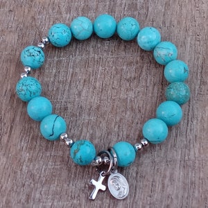 May include: A turquoise beaded bracelet with a silver charm featuring a cross and a depiction of the Virgin Mary.