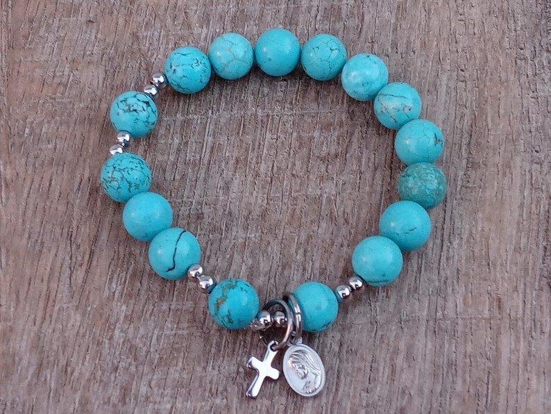 May include: A turquoise beaded bracelet with a silver charm featuring a cross and a depiction of the Virgin Mary.