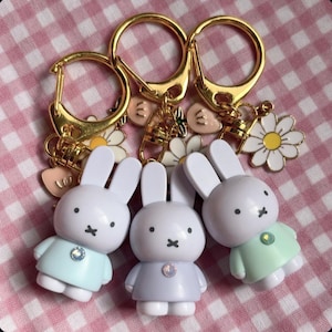 May include: Three keychains with gold-colored rings and charms. Each keychain features a white bunny figure wearing a pastel-colored dress. The charms include daisies and heart-shaped pendants. The background is a pink and white checkered pattern.