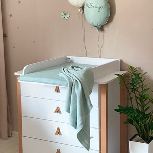 Puede incluir: Una cómoda blanca con cuatro cajones y una superficie para cambiar pañales. La superficie para cambiar pañales tiene una almohadilla para cambiar pañales de color azul claro y una manta de tejido de waffle de color azul claro. Dos globos de tela están en la pared detrás de la cómoda, uno blanco y otro verde claro con el nombre "M. Sonya" escrito en él. Hay pequeñas estrellas grises en la pared.