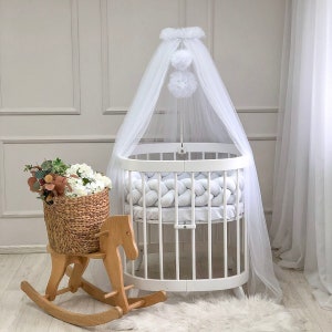 Peut inclure: Lit d'enfant rond blanc avec une canopée blanche et un pare-chocs tressé blanc. Un cheval à bascule en bois se trouve au premier plan.