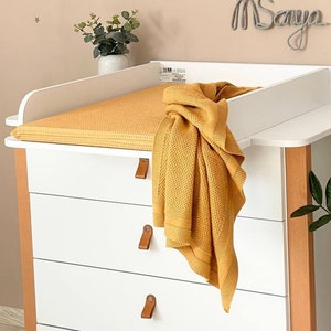 Puede incluir: Mesa cambiador blanca con cuatro cajones y una colchoneta para cambiar. La mesa cambiador tiene un marco de madera y una tapa blanca. Una manta amarilla está doblada en la colchoneta para cambiar. La mesa cambiador está decorada con una mariposa de madera y un letrero de nombre de alambre que dice "Msanya".