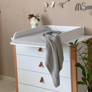Puede incluir: Mesa cambiadora blanca con cuatro cajones y una colchoneta cambiadora blanca. La mesa cambiadora tiene lados de madera marrón y tiradores de cuero marrón. Una manta de punto gris está drapeada sobre la colchoneta cambiadora. La pared detrás de la mesa cambiadora es de color beige claro con estrellas grises y un móvil de cometa rosa y beige con el nombre "Saya" escrito en cursiva.