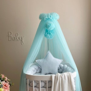 May include: A baby crib with a light blue canopy and star-shaped pillow. The crib has a white frame with a braided bumper and a white blanket. The word "Baby" is written on the wall in cursive.