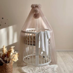 Può includere: Un lettino rotondo bianco con un materasso bianco e un paraurti intrecciato beige. Un asciugamano bianco è appeso sul lato del lettino. Un baldacchino in tulle rosa con pompon è drappeggiato sul lettino.