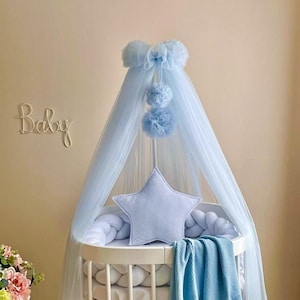 Può includere: Un baldacchino per culla per neonati di colore azzurro chiaro con un cuscino a forma di stella e un paraurti intrecciato bianco. Il baldacchino ha un bordo superiore arricciato e tre pompon azzurri chiari che pendono. La parola "Baby" è scritta sul muro dietro la culla.