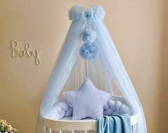 May include: A light blue baby crib canopy with a star-shaped pillow and a white braided bumper. The canopy has a ruffled top and three light blue pom-poms hanging from it. The word "Baby" is written on the wall behind the crib.