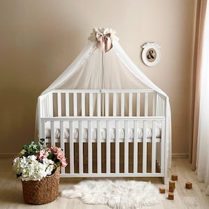 May include: White baby crib with a white canopy and a pink bow. The crib has a white mattress and a white sheet. There are wooden blocks on the floor next to the crib.