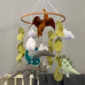 May include: A baby mobile with a green, blue, and gray dinosaur, a brown pterodactyl, and a gray mountain. The mobile is made of felt and has a wooden ring.