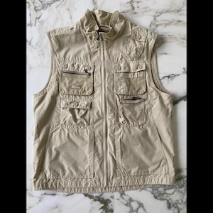Peut inclure: Un gilet sans manches beige avec plusieurs poches et une fermeture éclair sur le devant. Le gilet a un col et plusieurs poches avec fermetures éclair et boutons. Le gilet est en matière légère.
