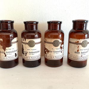 Peut inclure: Quatre flacons de médicaments anciens en verre brun avec des étiquettes. Les étiquettes indiquent le texte "Acidum Acetylsalicylicum" et "E.R. Squibb & Sons, New York". Les flacons sont de tailles variables et ont des couvercles brun foncé.