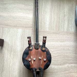 May include: A traditional stringed instrument with a round, dark brown body and a long, slender neck. The body features a decorative pattern with cowrie shells. The instrument has a dark brown finish and is likely handmade.