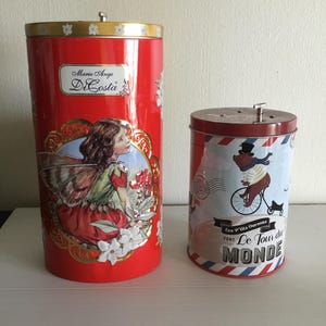 May include: Two red metal music boxes. The first music box has a fairy with wings and flowers on it. The second music box has a bear riding a bicycle with the text "Le Tour du Monde" on it.