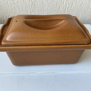 Könnte beinhalten: Eine braune Keramik-Auflaufform mit Deckel. Die Form ist rechteckig mit abgerundeten Ecken und einer gesprenkelten Oberfläche. Der Deckel hat einen gebogenen Griff und ein kleines Entlüftungsloch. Die Form steht auf einer weißen Oberfläche.