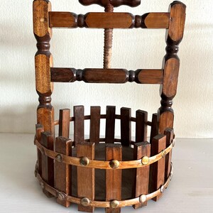 Può includere: Un'antica pressa per bottiglie di vino in legno con un meccanismo a vite e una piccola maniglia in legno. La pressa ha un cestello cilindrico fatto di doghe di legno verticali ed è tenuto insieme da fasce e rivetti metallici. Il legno è di colore marrone caldo.