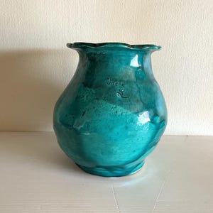Può includere: Un vaso in ceramica turchese con bordo smerlato. Il vaso ha un corpo bulboso e una superficie strutturata. Il nome "Irene" è inciso sul lato. Il vaso è alto circa 25 cm.
