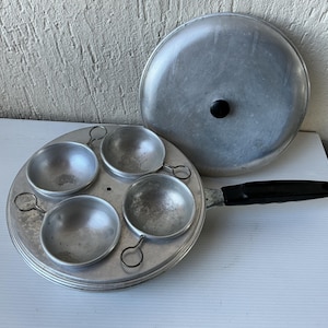 Può includere: Una vecchia poachiera per uova in alluminio con coperchio. La poachiera ha quattro tazze individuali e un lungo manico nero. Il coperchio ha un pomello nero. La poachiera è su una superficie bianca.