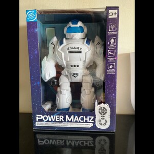 Könnte beinhalten: Weiß-blauer Power Machz Roboter-Spielzeug in einer Box. Der Roboter hat das Wort "SMART" auf der Brust und "HIDE" auf der Taille. Die Box enthält eine Fernbedienung und den Text "POWER MACHZ".